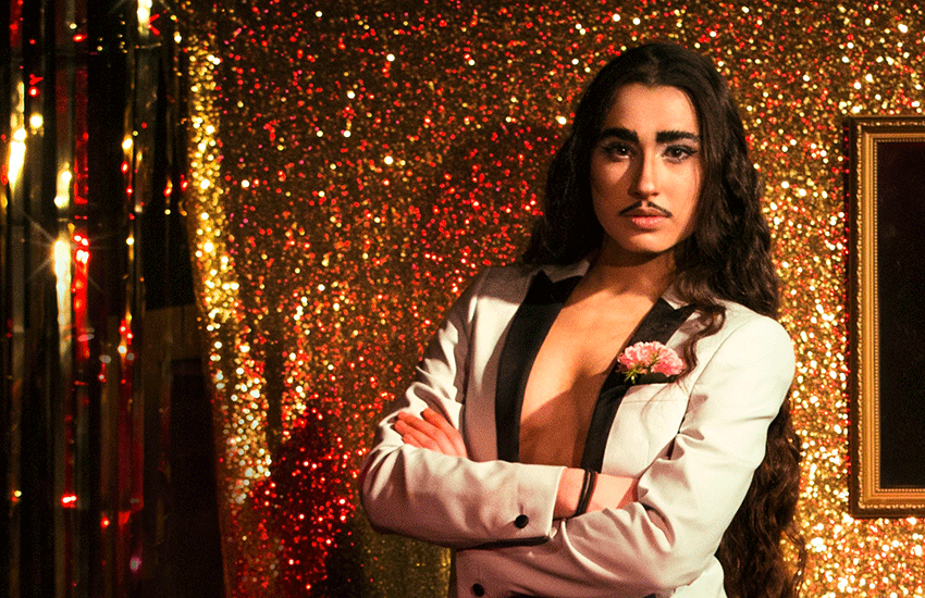 Imagen de la drag king christian adore posando con una chaqueta blanca formal frente a un fondo con una tela llena de glitter.